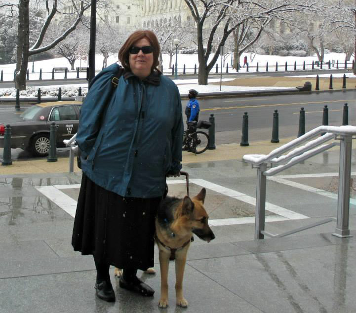 Photo of a woman with a German Shepherd guide dog on a city sidewalk on a snowy day