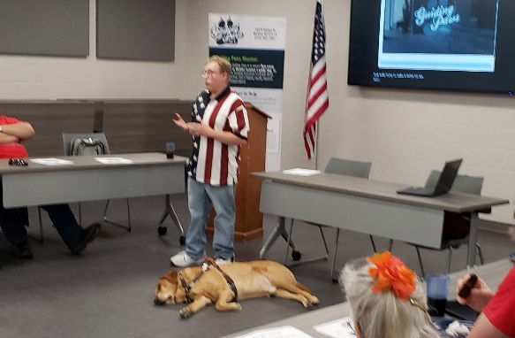 photo of Andrew speaking to a group while his guide dog Orzo lies at his feet