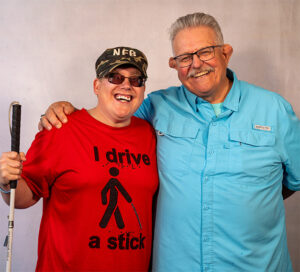 photo of Andrew Crane at the NFB National Convention 2024, holding his cane & wearing a t-shirt that says "I drive a stick," standing beside his father Dave